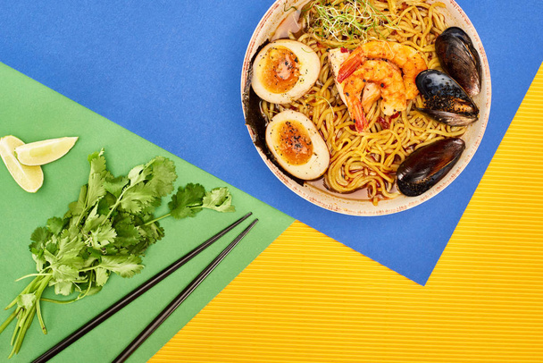 вид на пряный рамен из морепродуктов рядом со свежими ингредиентами и палочками для еды на разноцветной поверхности
 - Фото, изображение