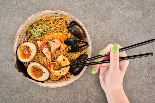 cropped view of woman holding chopsticks near spicy seafood ramen on grey surface - Fotografie, Obrázek