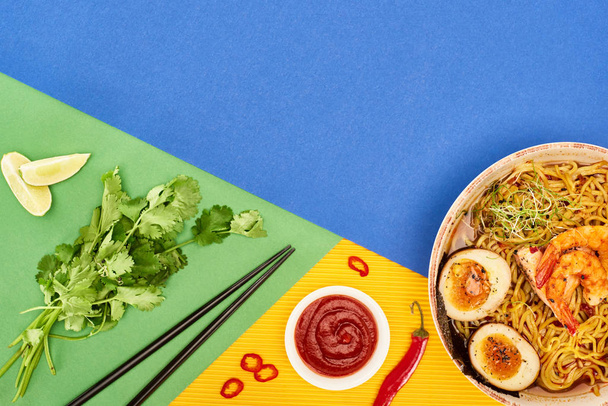 top view πικάντικα θαλασσινά ράμεν κοντά σε φρέσκα υλικά, σάλτσα τσίλι και chopsticks στην πολύχρωμη επιφάνεια - Φωτογραφία, εικόνα
