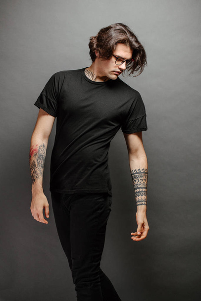 Hipster handsome male model with glasses wearing black blank t-shirt and black jeans with space for your logo or design in casual urban style - Foto, immagini