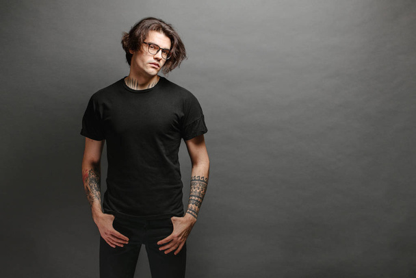 Hipster handsome male model with glasses wearing black blank t-shirt and black jeans with space for your logo or design in casual urban style - 写真・画像
