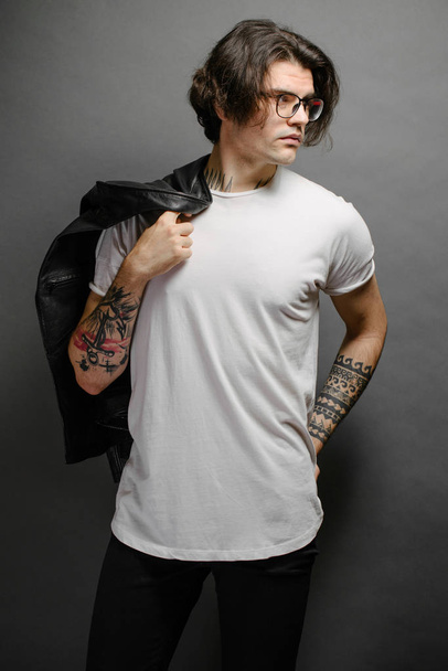 Hipster όμορφο ανδρικό μοντέλο με γυαλιά που φορούν λευκό κενό t-shirt και μαύρο τζιν με χώρο για το λογότυπο ή το σχέδιό σας σε casual urban στυλ - Φωτογραφία, εικόνα
