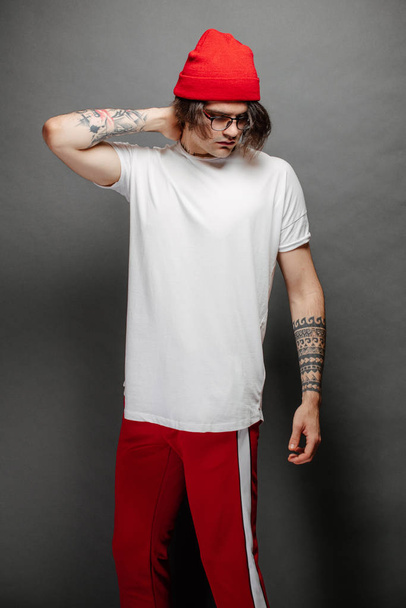 Hipster handsome male model with glasses wearing white blank t-shirt and red pants and a red hat with space for your logo or design over gray background. - Zdjęcie, obraz