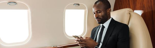 πανοραμική λήψη του όμορφου Αφροαμερικανού επιχειρηματία που χρησιμοποιεί smartphone σε ιδιωτικό τζετ  - Φωτογραφία, εικόνα