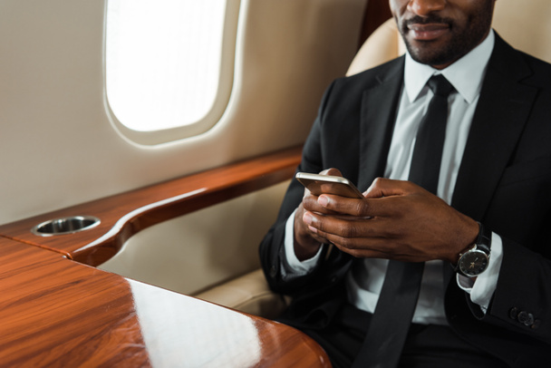 καλλιεργημένη άποψη του Αφροαμερικανού επιχειρηματία με κοστούμι χρησιμοποιώντας smartphone σε ιδιωτικό τζετ  - Φωτογραφία, εικόνα