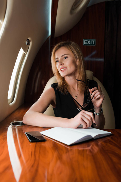 selektivní zaměření veselá podnikatelka drží pero a brýle v blízkosti notebooku a smartphone s prázdnou obrazovkou  - Fotografie, Obrázek