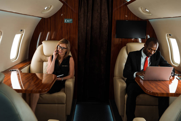 Afrikaans Amerikaans zakenman met behulp van laptop in de buurt van zakenvrouw praten op smartphone in prive-jet  - Foto, afbeelding