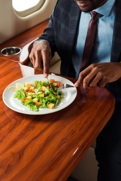 cropped view of african american businessman holding cutlery near salad in private jet  - Фото, изображение