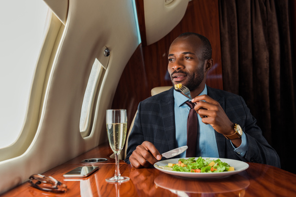 handsome african american man eating salad in private jet  - Foto, afbeelding