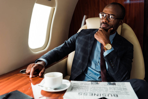 selective focus of african american businessman in glasses near business newspaper and cup in private plane  - Photo, Image