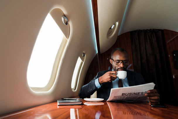 Αφροαμερικάνος επιχειρηματίας διαβάζει επιχειρηματικές εφημερίδες πίνοντας καφέ σε ιδιωτικό αεροπλάνο  - Φωτογραφία, εικόνα