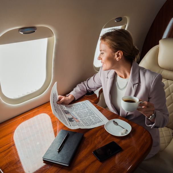 happy businesswoman holding cup and business newspaper near smartphone with blank screen in private plane  - Fotografie, Obrázek