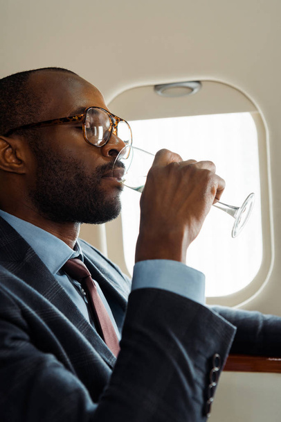 handsome african american businessman in glasses drinking champagne in private jet  - Photo, Image