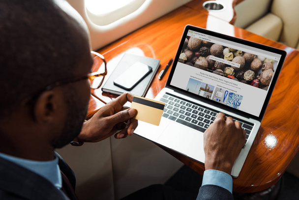 selectieve focus van Afrikaanse Amerikaanse zakenman met creditcard in de buurt van laptop met depositfoto 's website in prive-vliegtuig  - Foto, afbeelding