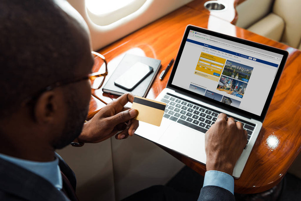 selective focus of african american businessman holding credit card near laptop with booking website in private plane  - Фото, изображение