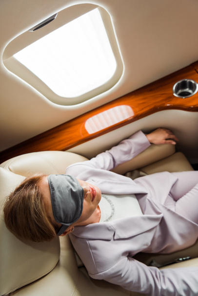 top view of businesswoman with sleeping mask sitting near airplane window in private plane  - Fotografie, Obrázek