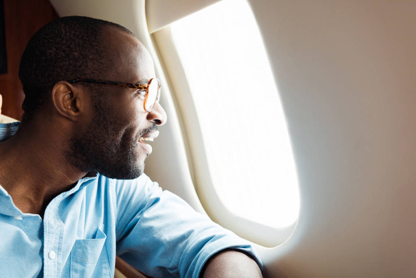 Χαρούμενος Αφροαμερικάνος με γυαλιά κοιτάζοντας το παράθυρο του αεροπλάνου.  - Φωτογραφία, εικόνα