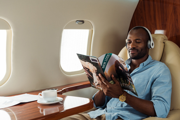 アフリカ系アメリカ人男性の選択的焦点は民間航空機での無線ヘッドフォン読み取り雑誌  - 写真・画像