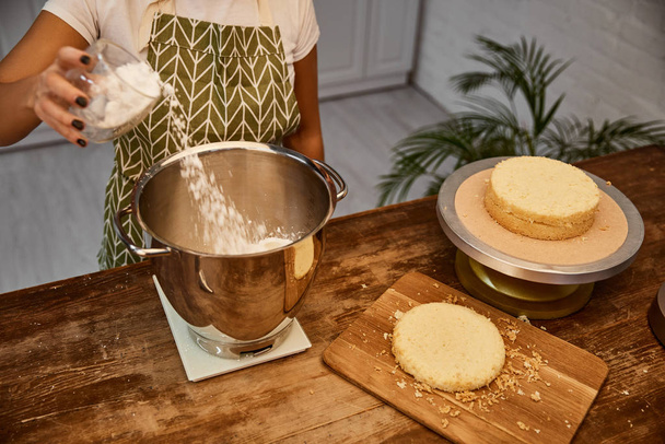 Vue recadrée du confiseur ajoutant de la farine dans un bol à côté des couches de gâteau sur la table
 - Photo, image