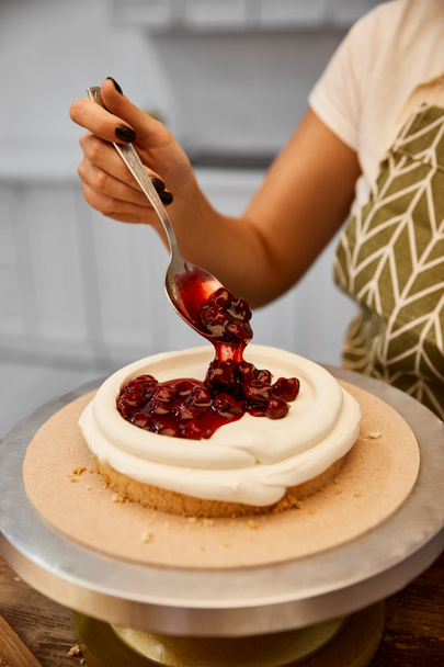 Vista recortada del confitero poniendo mermelada de bayas en la capa de pastel con crema
 - Foto, imagen