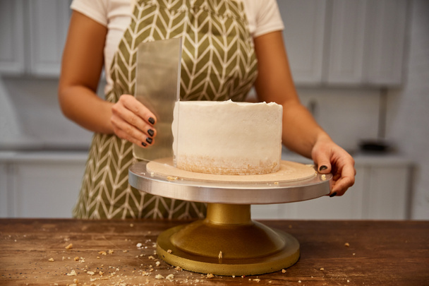 Vista recortada de la crema alineadora confitera femenina con raspador en la torta de esponja
 - Foto, imagen