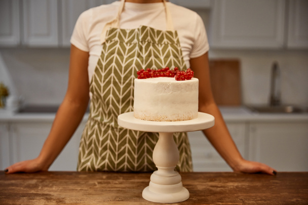 Vista recortada del confitero de pie junto a la torta sabrosa con grosella roja en la mesa
 - Foto, imagen