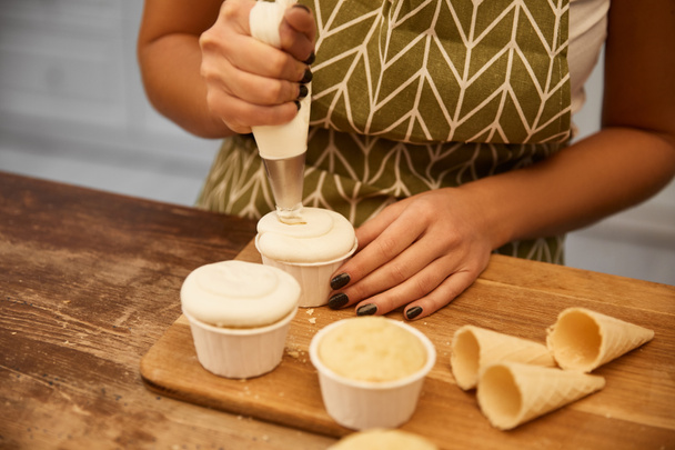 Καλλιεργημένη άποψη του ζαχαροπλάστη προσθέτοντας νόστιμη κρέμα σε ψημένα cupcakes  - Φωτογραφία, εικόνα