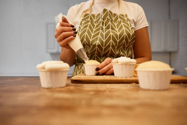 Selective focus of confectioner adding sweet cream on cupcakes on wooden table, cropped view - Foto, immagini