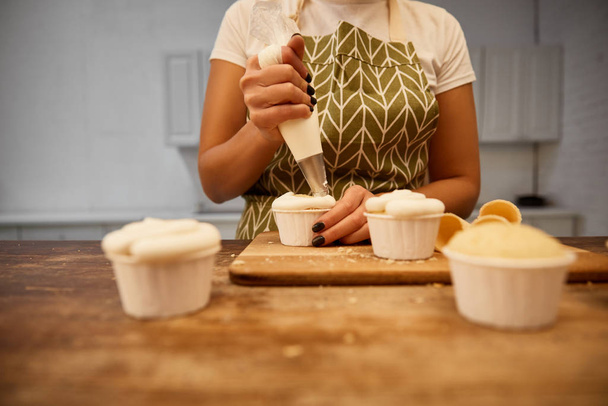 Gekropte weergave van banketbakker met banketbakkerszak gieten crème op cupcakes - Foto, afbeelding