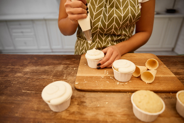 Gekropte weergave van banketbakker gieten crème op cupcakes naast wafelkegels op snijplank - Foto, afbeelding