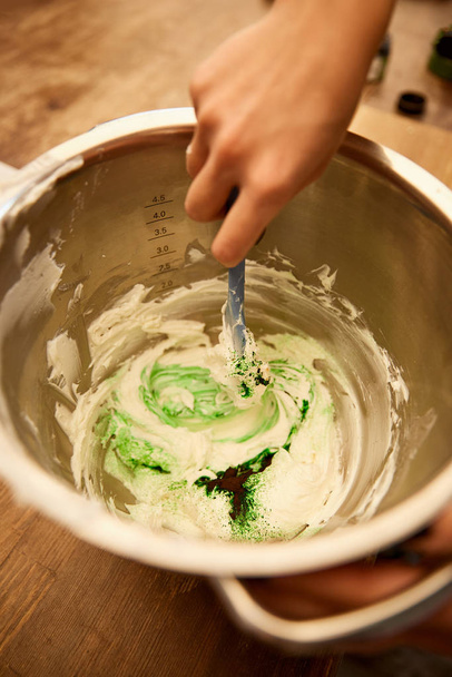 abgeschnittene Ansicht des Konditors, der Sahne mit grüner Lebensmittelfarbe in Schüssel mixt - Foto, Bild