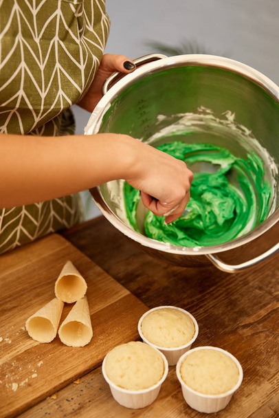 Обрезанный вид кондитера, смешивающего сладкий крем с кексами и вафельными рожками на столе
 - Фото, изображение
