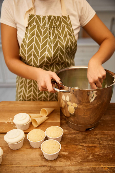 Cropped view of confectioner making cream beside cupcakes and waffle cones on wooden table - Photo, Image