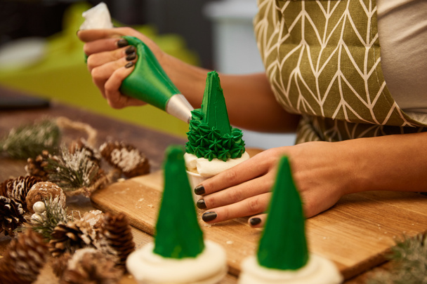 Selective focus of confectioner decorating tasty Christmas tree cupcakes beside spruce cones on table, cropped view  - Photo, Image
