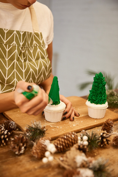 Vista recortada del pastelero usando una bolsa de pastelería con crema en los cupcakes de árbol de Navidad junto a los conos de abeto en la mesa
 - Foto, Imagen