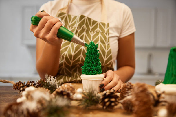 Зіпсований вид кондитера прикрашає різдвяний кекс зеленим кремом поруч шишок з ялинкою на столі. - Фото, зображення