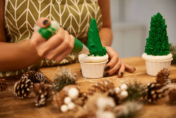 Vista recortada del confitero haciendo cupcakes de árbol de Navidad con crema verde con conos de abeto en la mesa
 - Foto, imagen
