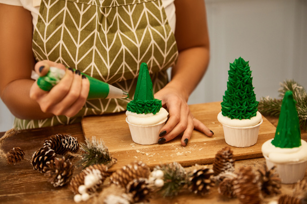 Vista cortada de confeiteiro fazendo cupcakes de árvore de Natal com creme doce verde ao lado de cones de abeto na mesa
 - Foto, Imagem