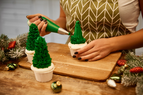 Oříznutý pohled cukráře dělat vánoční stromeček cupcakes vedle smrkových větví a vánoční koule na stole - Fotografie, Obrázek