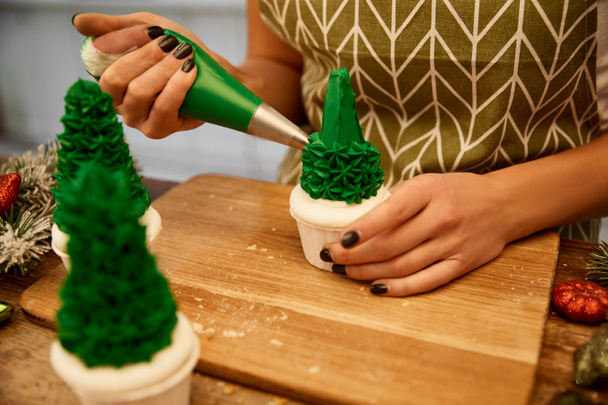 Vue recadrée du confiseur ajoutant de la crème sucrée sur les cupcakes de sapin de Noël avec des branches de pin et des boules de Noël sur la table
 - Photo, image