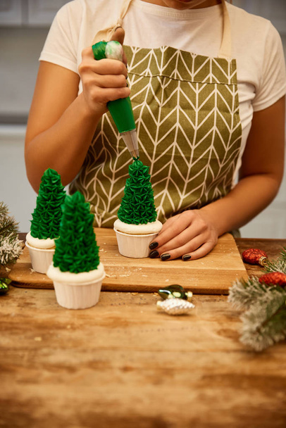 abgeschnittene Ansicht des Konditors Zugabe von Sahne auf Weihnachtsbaum Cupcakes mit Tannenzweigen und Weihnachtsdekor auf dem Tisch - Foto, Bild