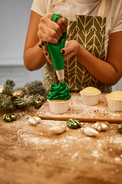Przycięty widok cukiernika podejmowania babeczki z zielonym kremem obok dekoracji świątecznej i gałązki sosny na stole - Zdjęcie, obraz