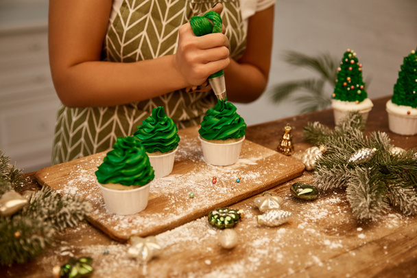 zugeschnittene Ansicht des Konditors, der Weihnachtsbaum-Cupcakes neben Fichtenzweigen und Weihnachtsschmuck auf dem Tisch dekoriert - Foto, Bild