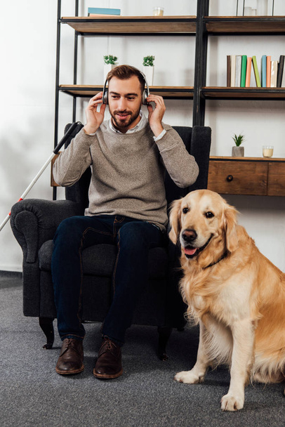 Visually impaired man listening music with headphones beside golden retriever at home - Foto, Imagen