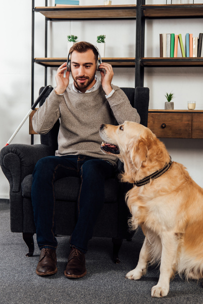 Blind man listening music with headphones beside golden retriever at home - Photo, Image