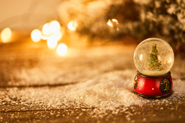 pequeña bola de nieve con árbol de Navidad de pie sobre una mesa de madera en la nieve con ramas de abeto y luces borrosas por la noche
  - Foto, Imagen