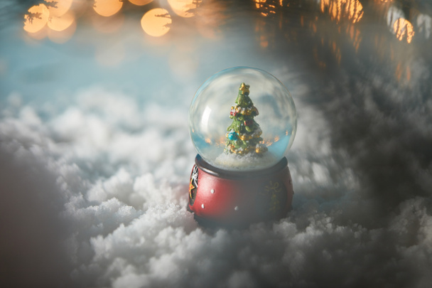 selective focus of decorative snowball with christmas tree standing on blue in snow with lights bokeh  - 写真・画像