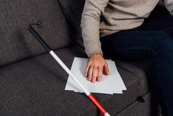 Cropped view of blind man reading braille font beside walking stick on sofa  - Photo, Image