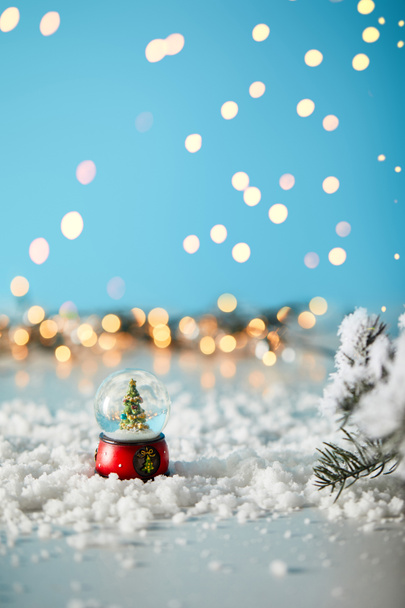 pequeño árbol de Navidad en bola de nieve de pie en azul con ramas de abeto en la nieve y luces borrosas
  - Foto, Imagen