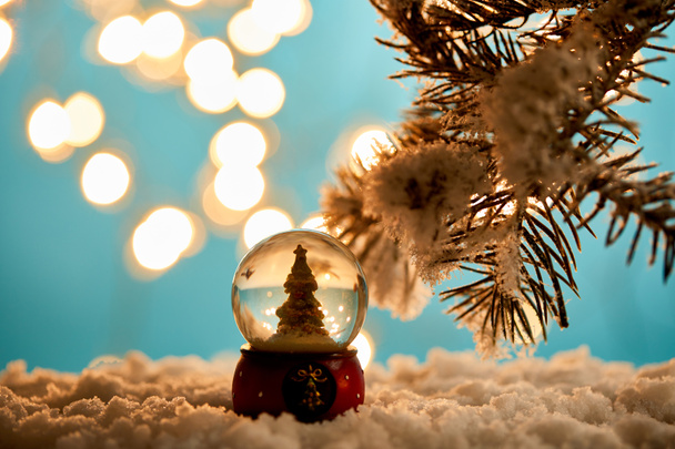 árbol de Navidad decorativo en bola de nieve de pie en azul con ramas de abeto en la nieve y luces borrosas
  - Foto, Imagen